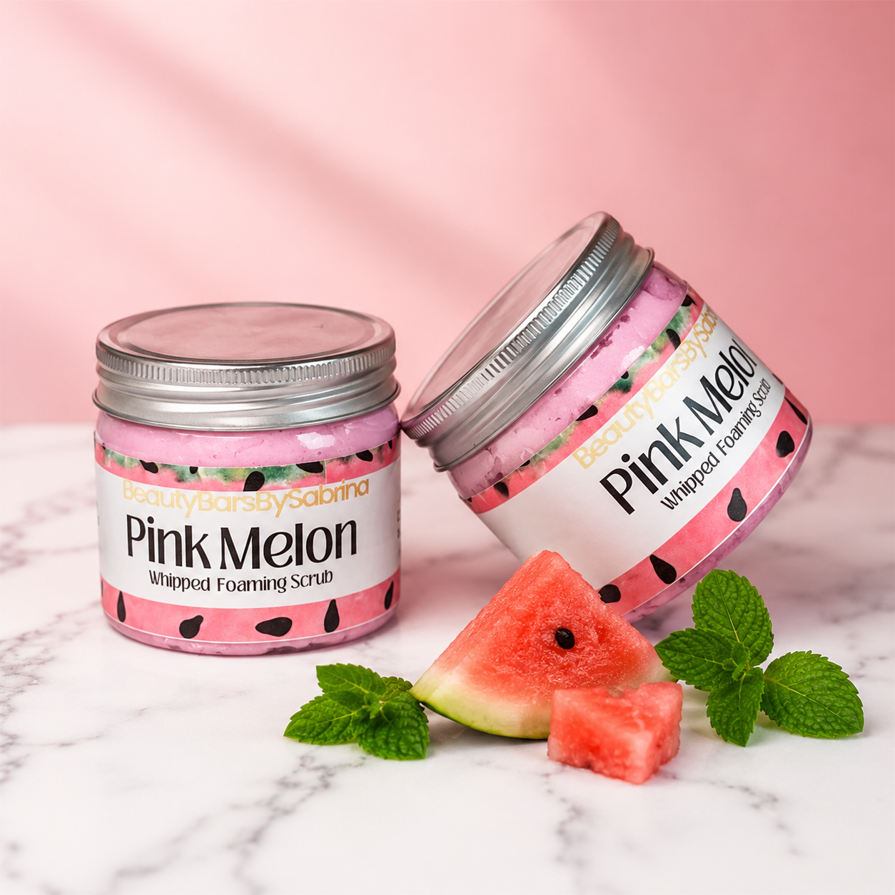 Two jars of Pink Melon whipped foaming scrub with watermelon slices and mint leaves on a marble surface.