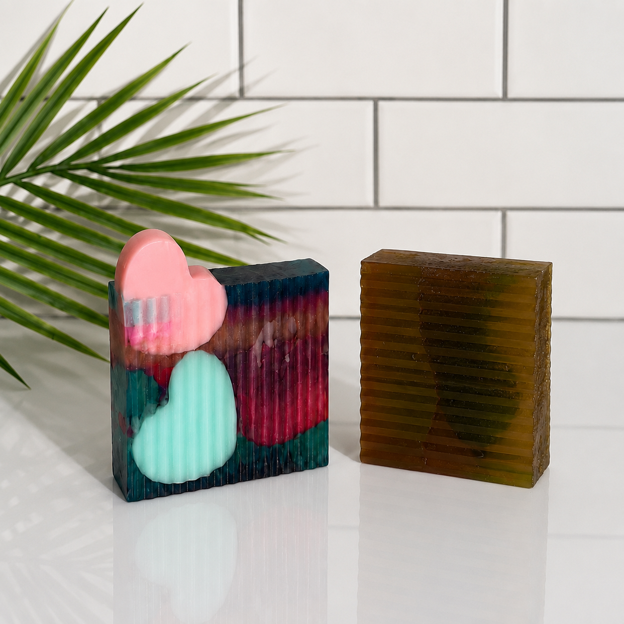 Two decorative rectangular bars of soap on a white surface against a white subway tile background, with a green palm leaf entering from the left. One soap is multicolor with pink and mint heart inlays, and the other is translucent olive-brown with horizontal ridges.