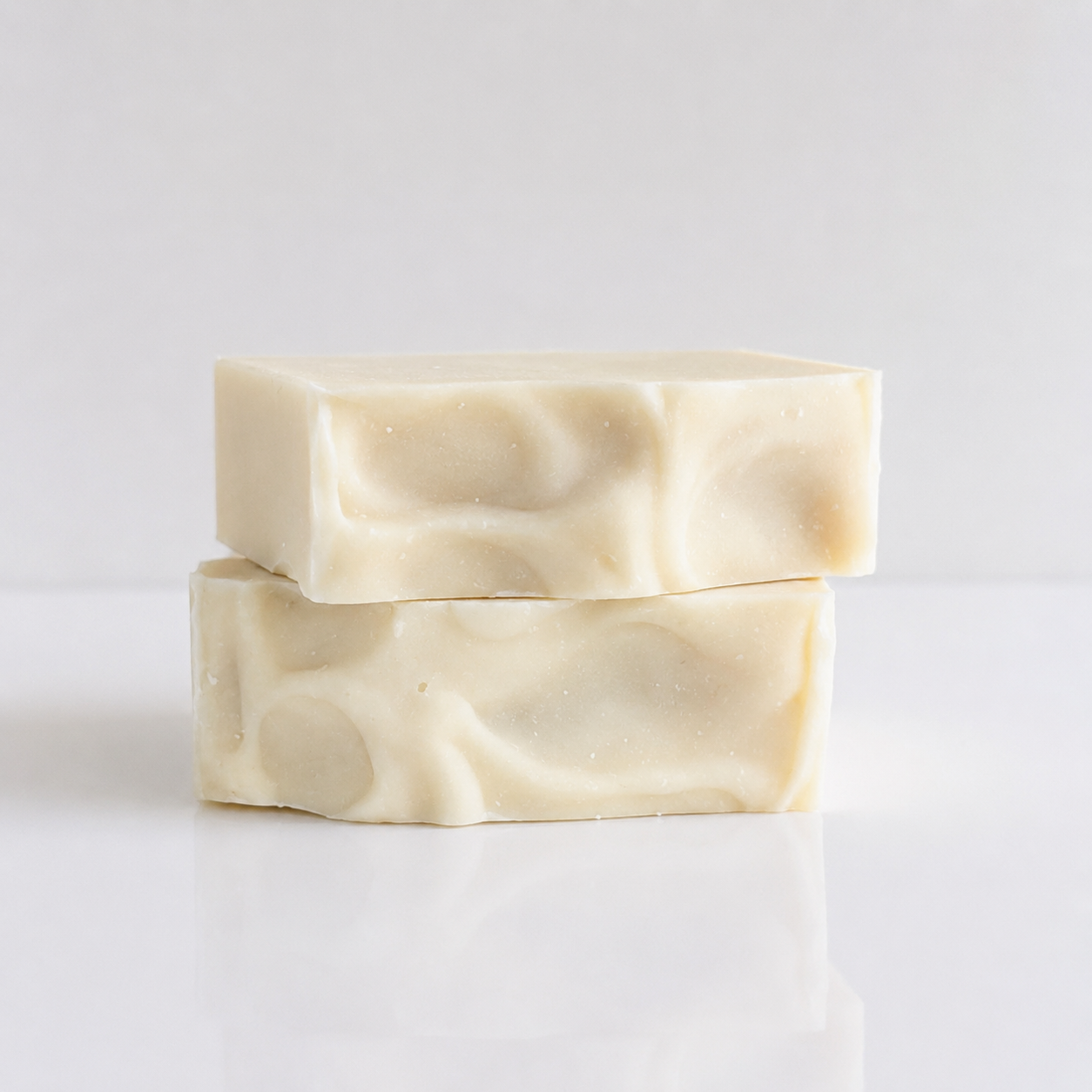 Two rectangular bars of handmade soap stacked on a white reflective surface against a light gray background.