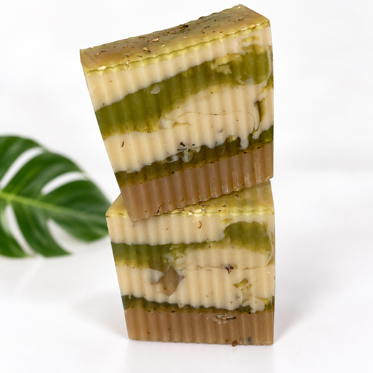 Two stacked rectangular bars of handmade soap with a marbled pattern of olive green, cream, and tan, set against a white background with a green monstera leaf on the left.