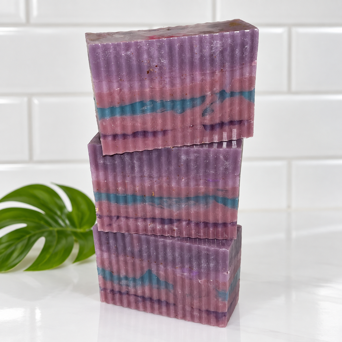Three stacked rectangular bars of handmade soap with a corrugated texture, featuring marbled layers of purple, pink, and teal blue, set against a white subway tile background with a green leaf prop on the left.