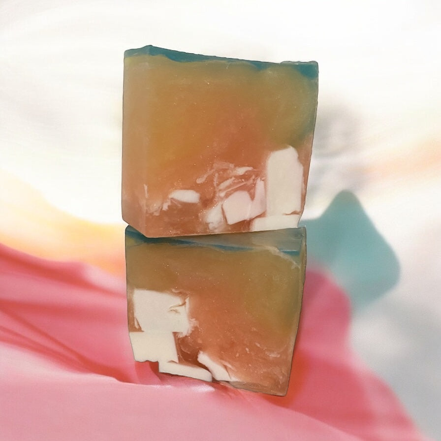 Two bars of soap with a translucent, candy-like appearance, possibly containing natural ingredients such as herbs or fruits, set against a soft-focused pink and blue background.