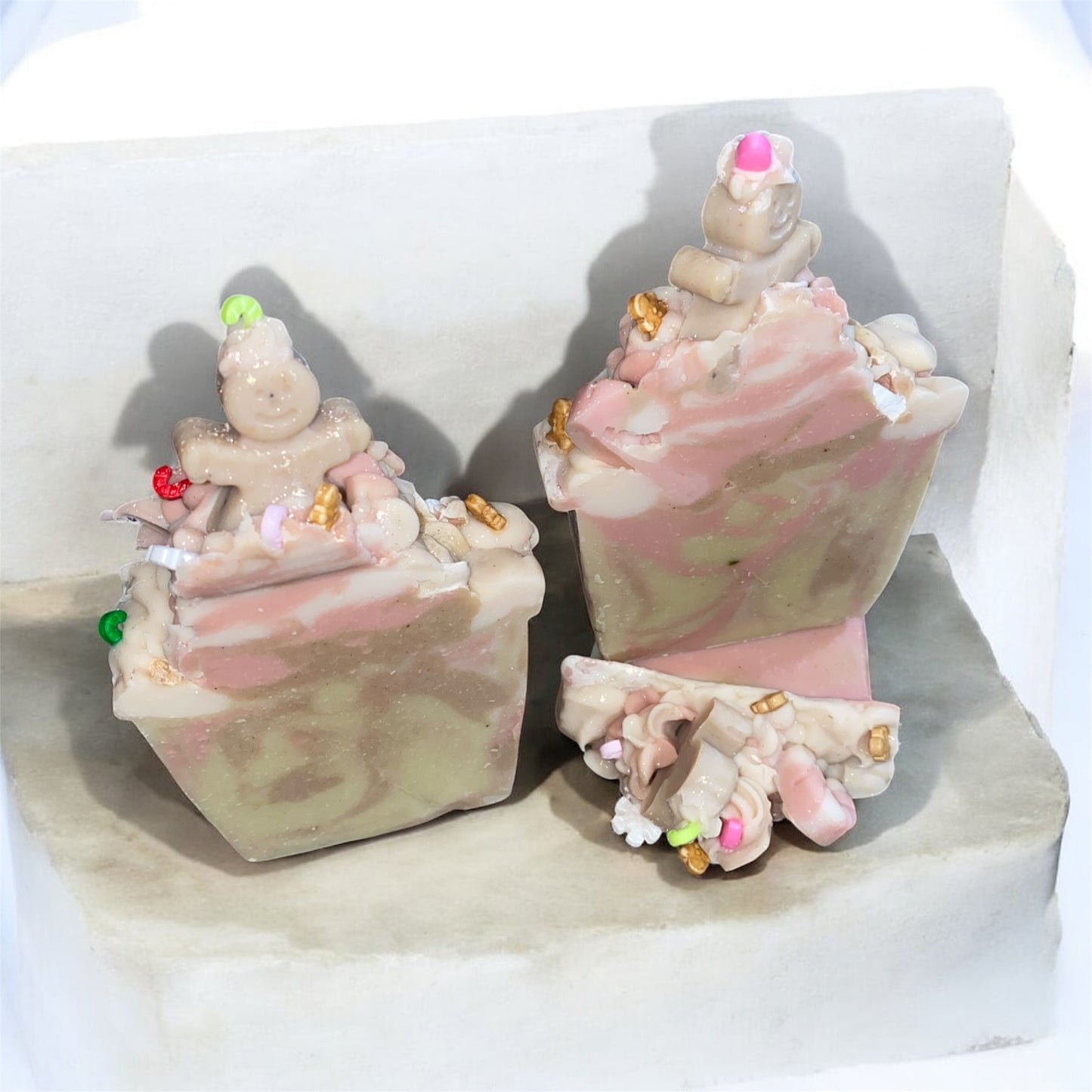 Two decorative soap resembling Christmas sprinkles, gingerbread man with pink and brown accents on a white surface.