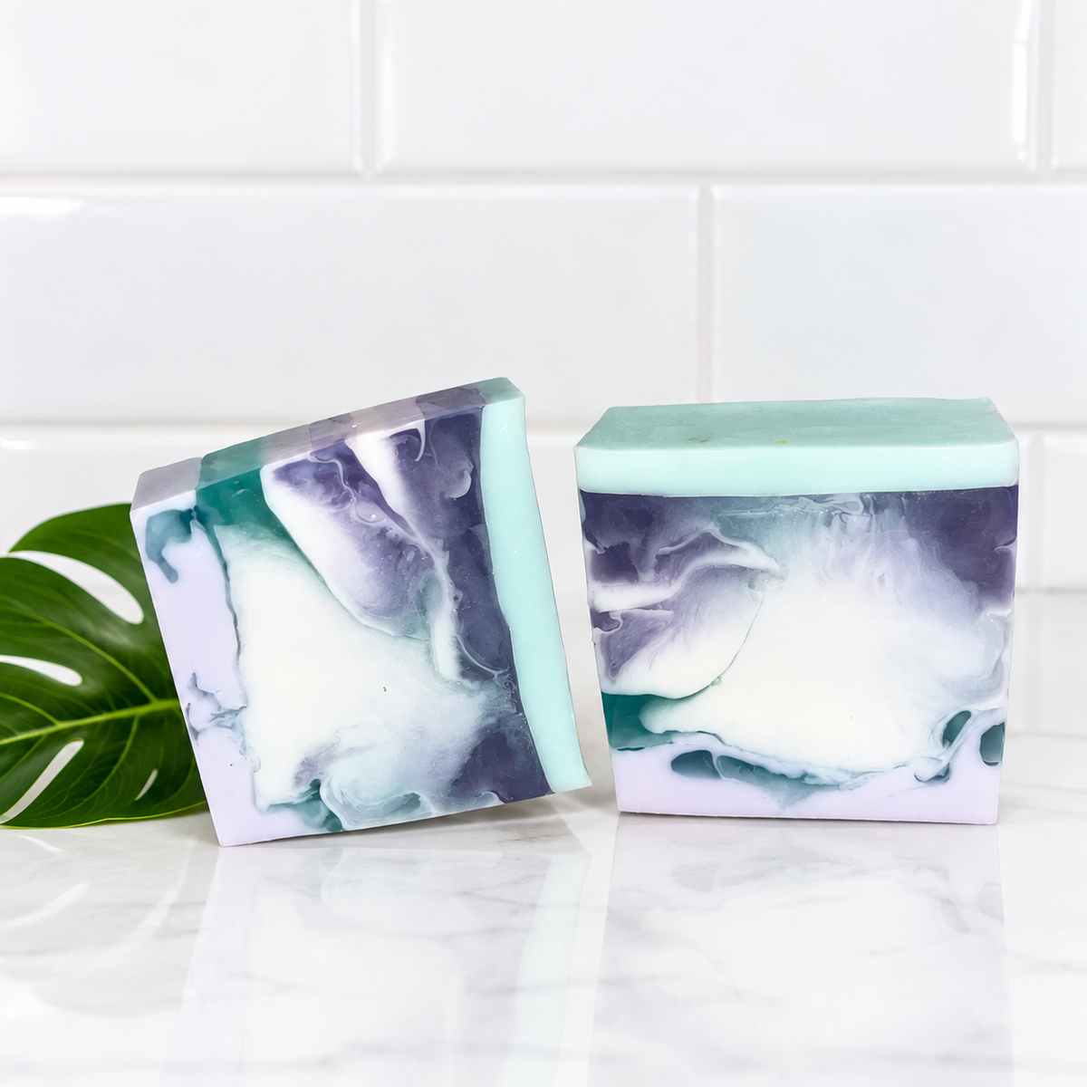 Two decorative rectangular bars of soap with a marbled swirl pattern in mint green, white, and purple, displayed on a white marble surface with a green leaf and a white subway tile background.