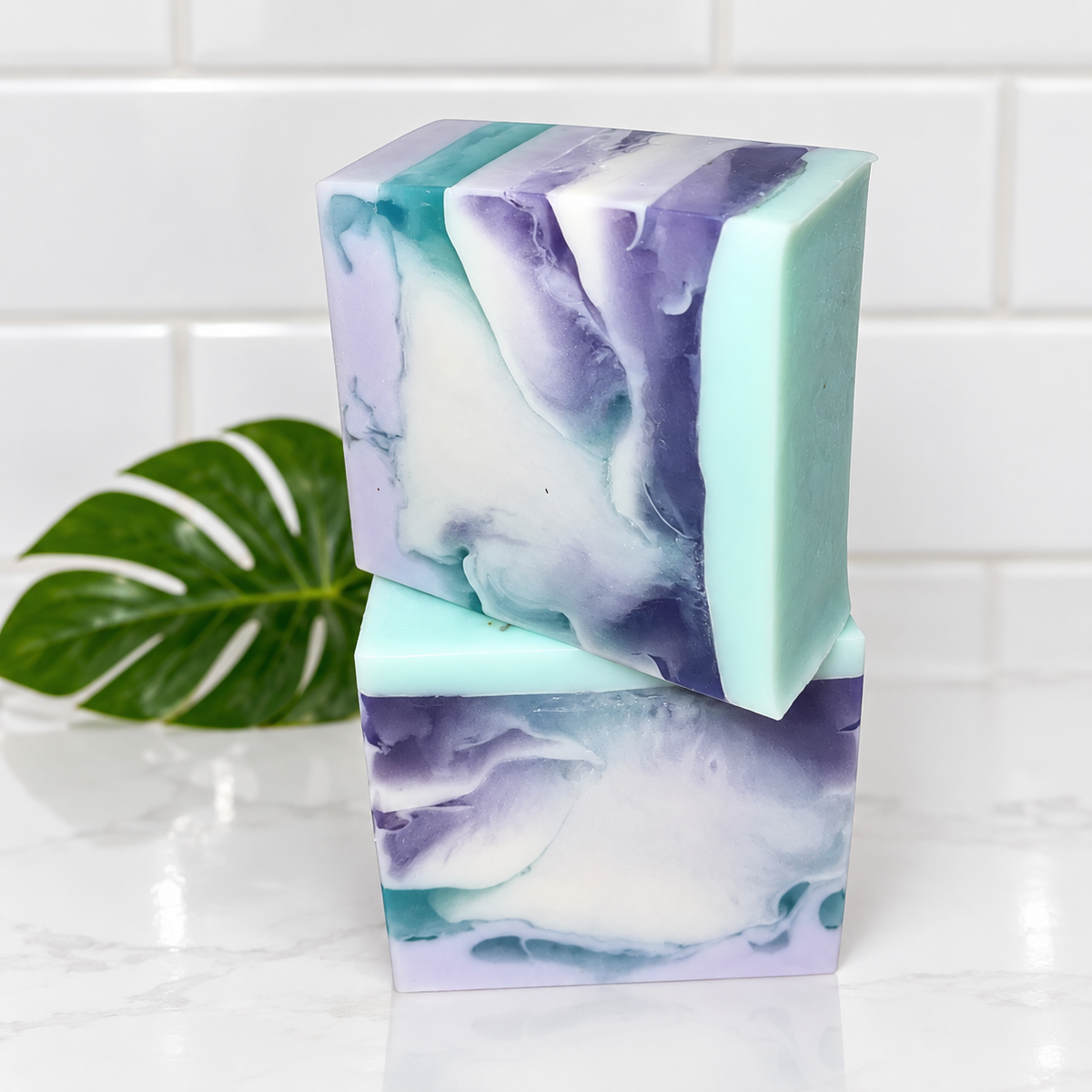 Two rectangular handmade soap bars stacked on a white marble surface, with a green monstera leaf in the background. The soaps have a marbled swirl pattern of mint green, purple, lavender, and white.