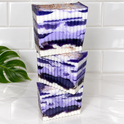 Three stacked rectangular bars of layered purple and white marbled soap with a brown clove on top, set on a white marble surface against a white subway tile background with a green leaf prop on the left.