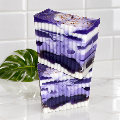 Two rectangular bars of layered purple and white soap with a marbled swirl pattern and a dusting of brown clove on top, standing on a white marble surface with a green leaf and white tiled background.