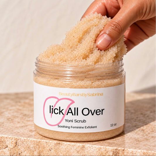 A hand scoops beige exfoliating scrub from a clear jar labeled 'lick All Over Yoni Scrub' on a stone surface with a neutral background.