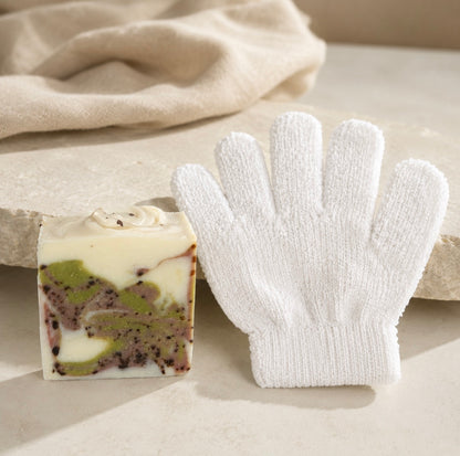 A white knit hand glove and a square decorative soap bar with cream, green, and dark berry swirls, styled on a light stone surface with a soft beige cloth in the background.