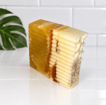A rectangular bar of handmade honey-colored and creamy white soap with visible layered flakes and small natural inclusions, photographed on a white tiled surface with a green leaf in the background.