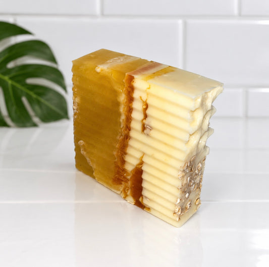 A rectangular bar of handmade honey-colored and creamy white soap with visible layered flakes and small natural inclusions, photographed on a white tiled surface with a green leaf in the background.
