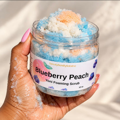 A hand with pink manicured nails holds a clear jar of Beauty Bars by Sabrina Blueberry Peach Yoni Foaming Scrub. The jar is filled with blue, white, and peach-colored foaming scrub, set against a soft, warm beige background.