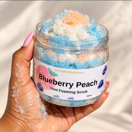 A hand with pink manicured nails holds a clear jar of Beauty Bars by Sabrina Blueberry Peach Yoni Foaming Scrub. The jar is filled with blue, white, and peach-colored foaming scrub, set against a soft, warm beige background.