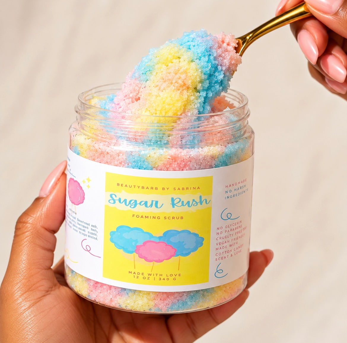 A hand holds a clear jar of colorful sugar scrub while another hand uses a gold spoon to scoop the pastel foam scrub. The label reads 'Sugar Rush Foaming Scrub' and shows the brand 'Beauty by Sabrina'.
