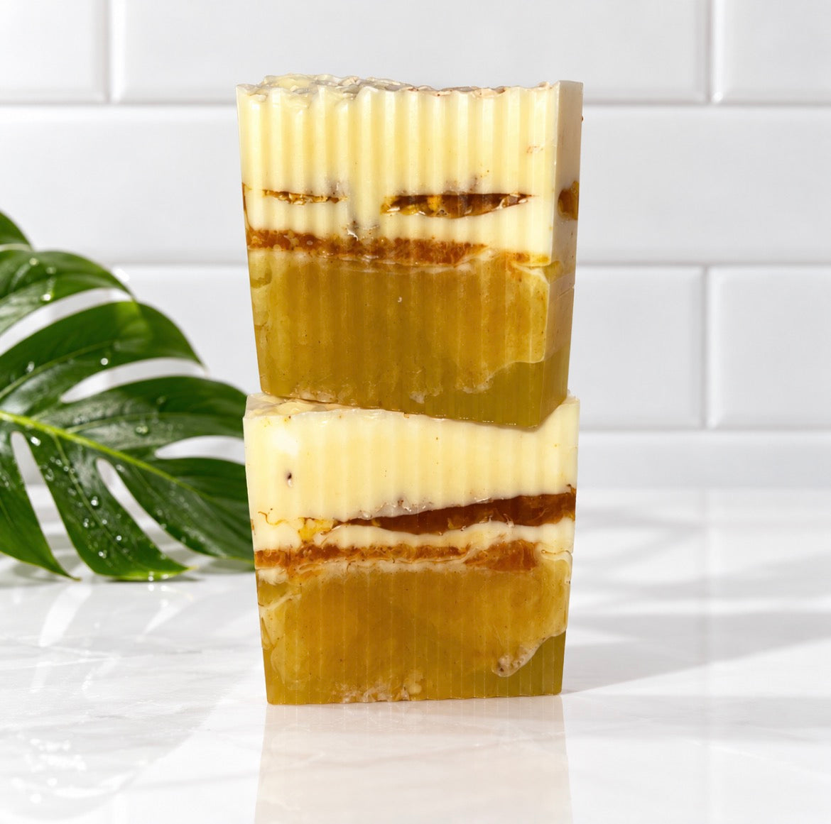 Two stacked rectangular bars of handmade soap with a layered design of creamy off-white, translucent olive green, and amber-brown, set against a white tiled background with a green leaf prop on the left.