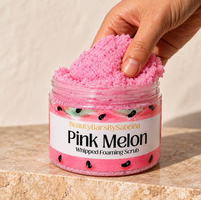 A hand scoops pink melon whipped foaming scrub from a clear plastic jar labeled 'BeautyBarsBySabrina Pink Melon Whipped Foaming Scrub' on a beige stone surface.