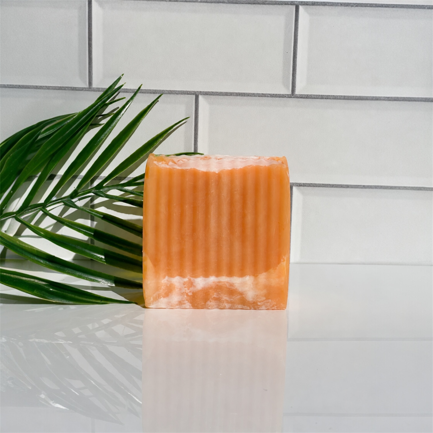 Orange soap bar on a white surface with a green leaf and tiled wall background