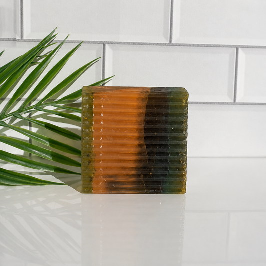A square soap with a ribbed texture, featuring a vertical color gradient from translucent orange on the left to dark green and green on the right, set against a white subway tile background with a green palm leaf on the left.