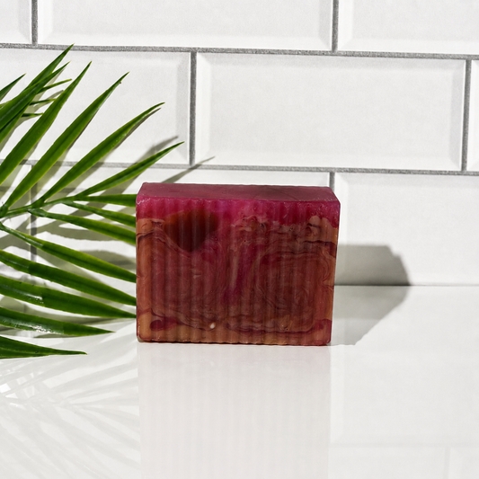 A rectangular bar of marbled magenta and reddish-brown soap or candle standing on a glossy white surface, with a green palm frond on the left and a white subway-tile wall behind it.