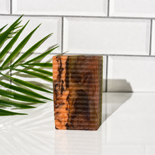 A rectangular soap block with a marbled pattern of orange, brown, and black, standing on a white surface in front of a white subway tile wall with a green palm leaf on the left.