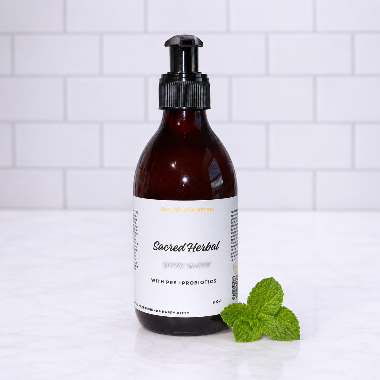 An amber glass pump bottle labeled 'Sacred Herbal Pure Water' with a black pump, set on a white marble surface with a white subway tile background and a small green mint leaf beside it.