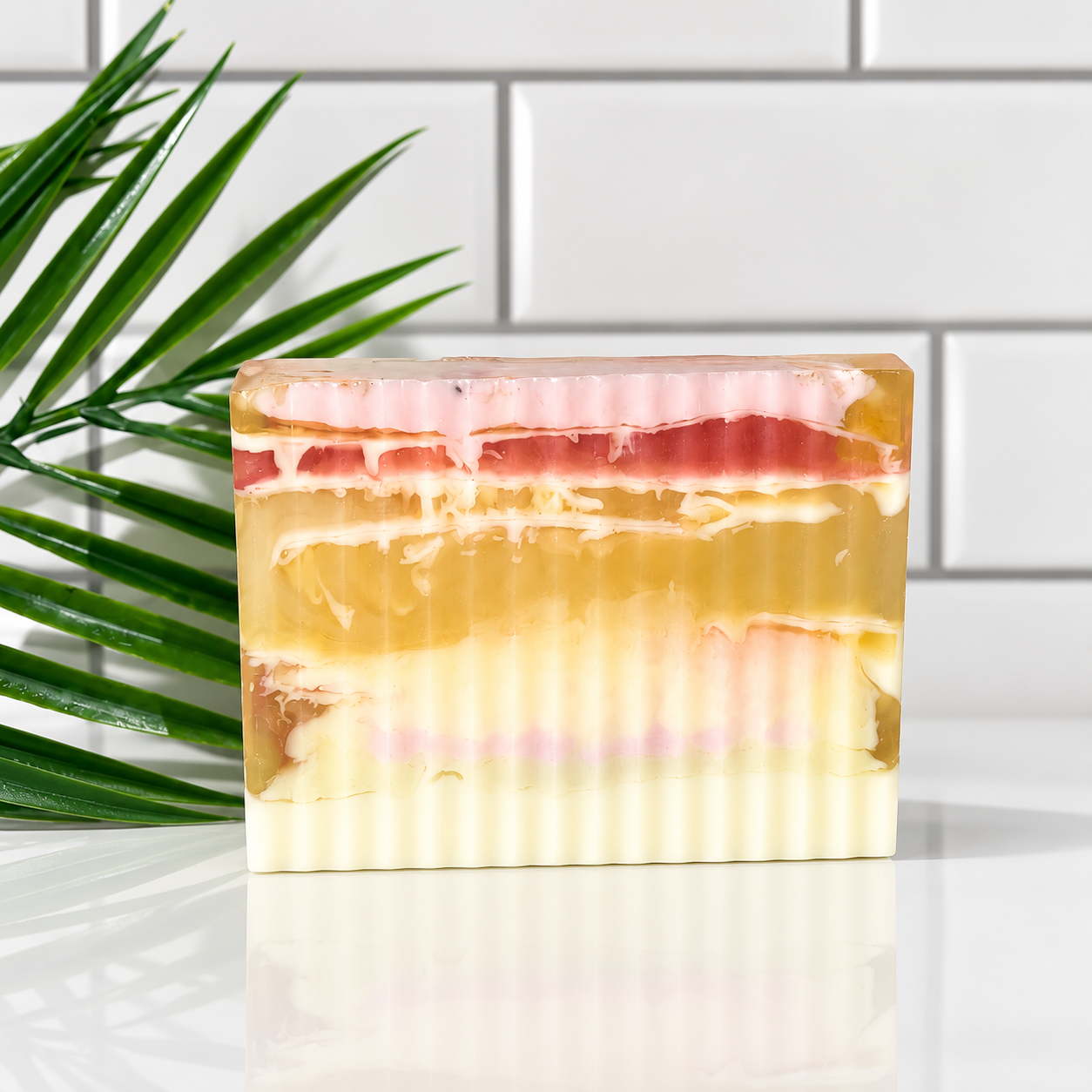 A rectangular bar of layered handmade soap with cream, yellow, pink, and red bands, standing on a reflective white surface with a green palm leaf on the left and a white subway tile wall in the background.