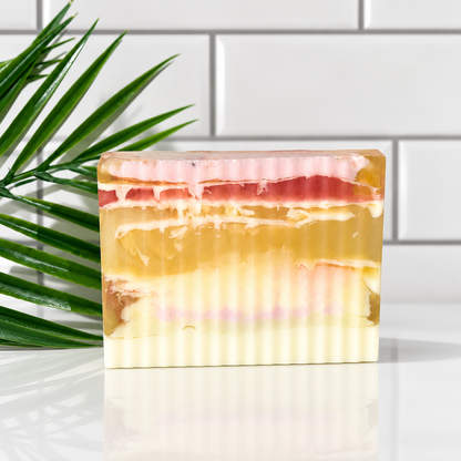 A rectangular bar of layered handmade soap with cream, yellow, pink, and red bands, standing on a reflective white surface with a green palm leaf on the left and a white subway tile wall in the background.
