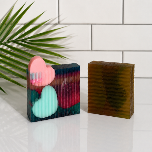 Two decorative rectangular bars of soap on a white surface against a white subway tile background, with a green palm leaf entering from the left. One soap is multicolor with pink and mint heart inlays, and the other is translucent olive-brown with horizontal ridges.
