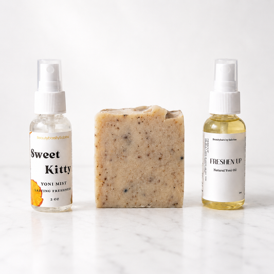 Three small products arranged on a white marble surface against a white background: a clear spray bottle labeled 'Sweet Kitty' on the left, a rectangular bar of speckled beige soap in the center, and a yellow oil spray bottle labeled 'FRESHEN UP' on the right.