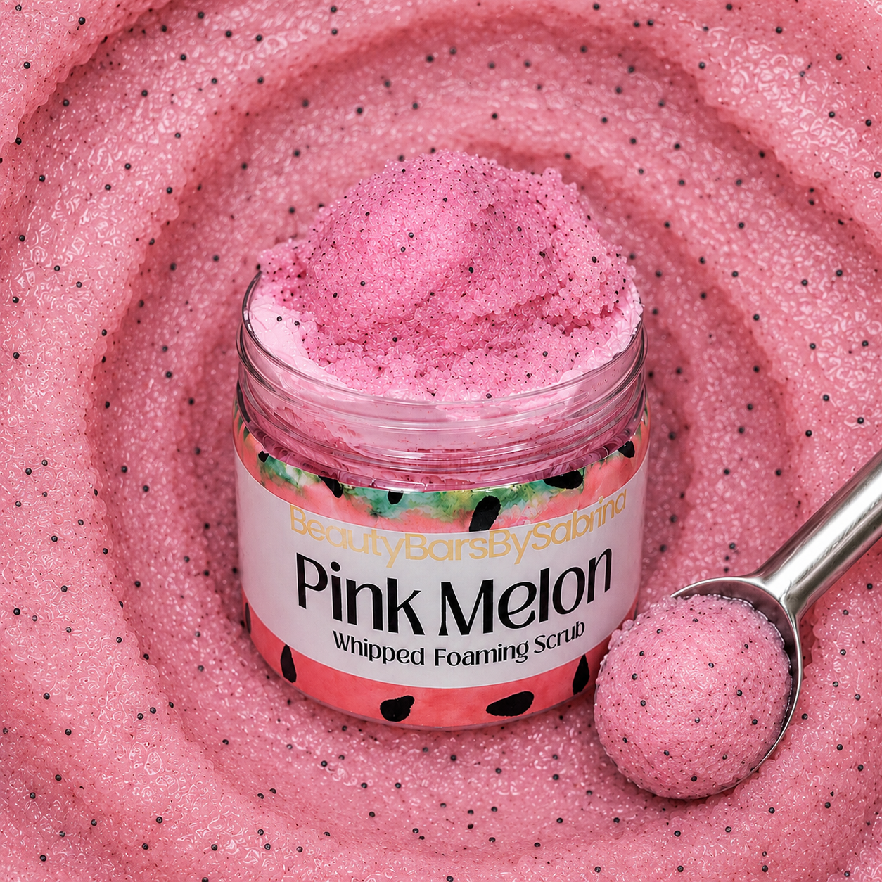 A jar of pink melon whipped foaming scrub by Beauty Bars by Sabrina, shown with a metal scoop holding a dollop of the scrub, set against a background of the same pink scrub texture.