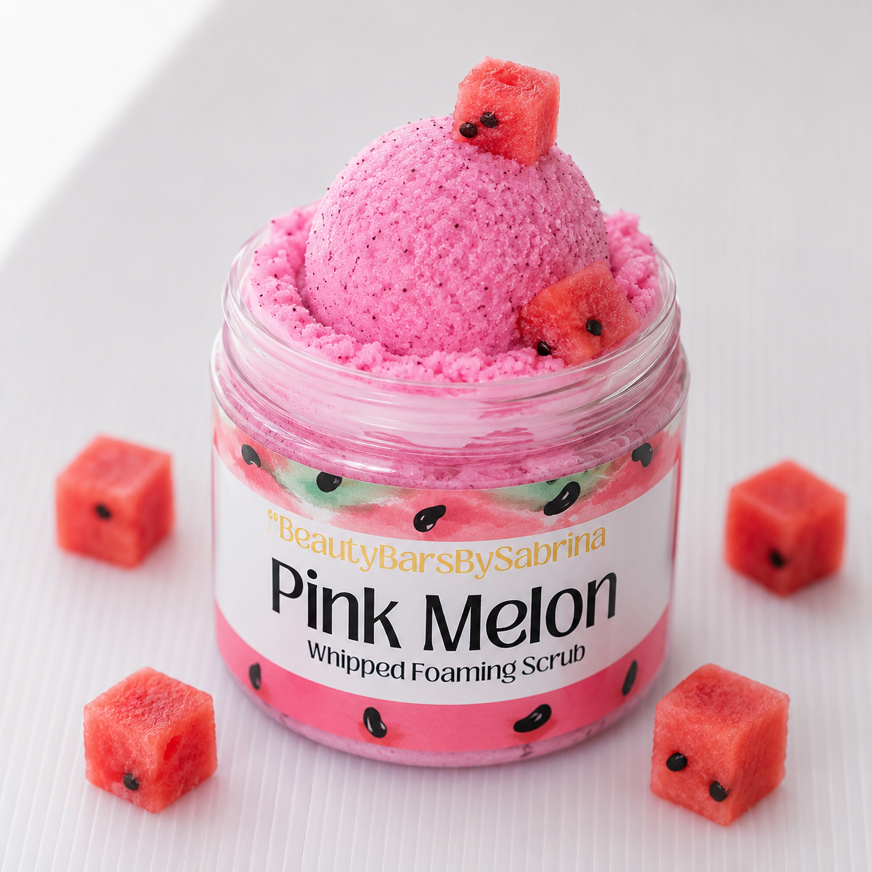 A clear jar of whipped foaming scrub labeled 'Pink Melon' by BeautyBarsBySabrina, filled with bright pink scrub and topped with watermelon pieces, with additional watermelon cubes arranged around it on a white surface.