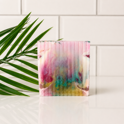 A rectangular soap with a colorful pattern in pink, teal, yellow, and white, standing on a white surface with a green palm leaf on the left and a white subway tile wall behind it.