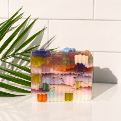 A rectangular handmade soap bar with colorful layered patterns and small gummy-like details on top, photographed on a white surface with a tiled wall and green palm leaves in the background.