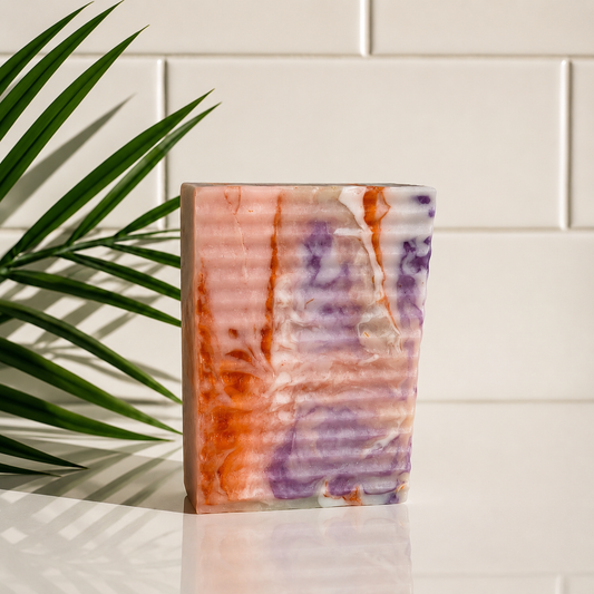 A rectangular bar of soap with a marbled pattern of orange, purple, and white, standing on a white surface in front of a white subway tile wall with a green palm leaf casting shadows.