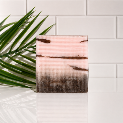 A rectangular bar of soap with horizontal pink, beige, and dark brown marbled stripes, standing on a reflective white surface with a green palm leaf on the left and a white subway tile wall in the background.
