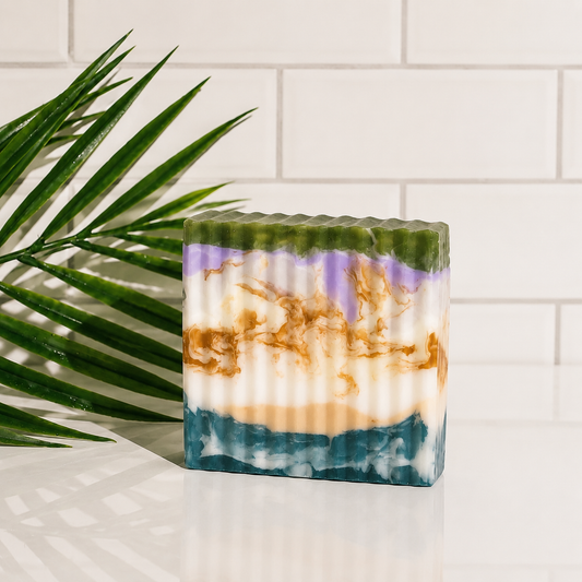 A rectangular marbled soap bar with green, purple, white, tan, and blue layers, styled on a white surface with a green palm leaf and a white subway tile background.