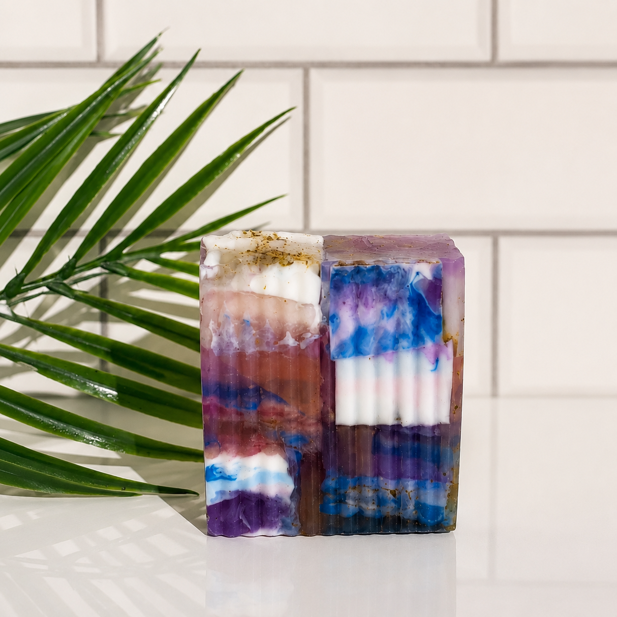 A handmade bar of soap with a marbled, layered pattern of purple, blue, white, and terracotta, standing on a white surface in front of a white subway tile wall with a green palm leaf on the left.
