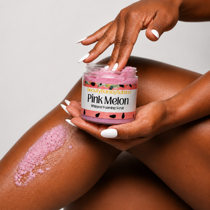 A jar of Pink Melon Whipped Foaming Scrub is held in hands with white manicured nails, with a smear of pink scrub visible on a person's leg against a light gray background.