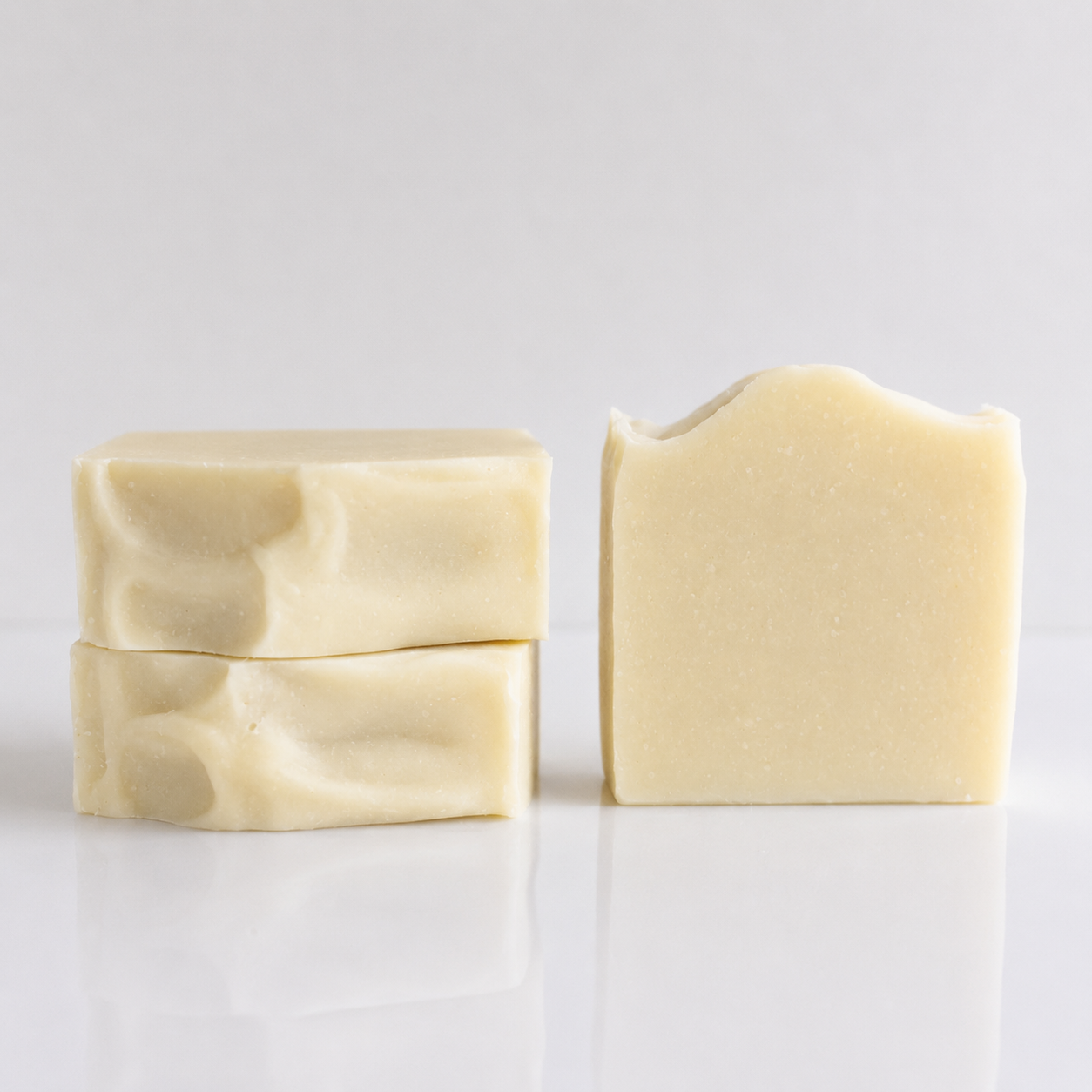 Three bars of handmade soap in a warm cream color, arranged on a reflective white surface against a light gray background.