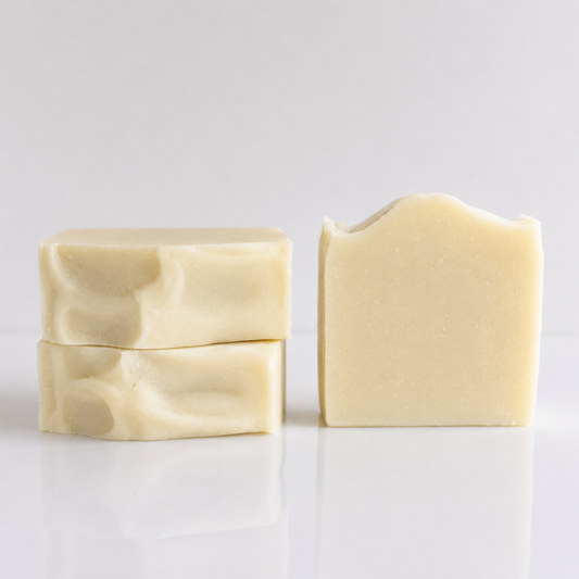 Three bars of handmade soap in a warm cream color, arranged on a reflective white surface against a light gray background.