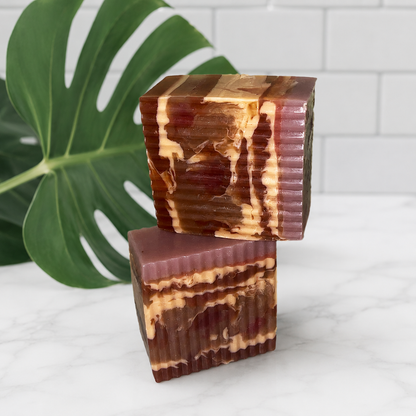Two handmade rectangular bar soaps stacked on a white marble surface, featuring a marbled pattern of deep reddish-brown, dark brown, and cream swirls. A large green monstera leaf is visible on the left, and a white subway tile backsplash is in the background.