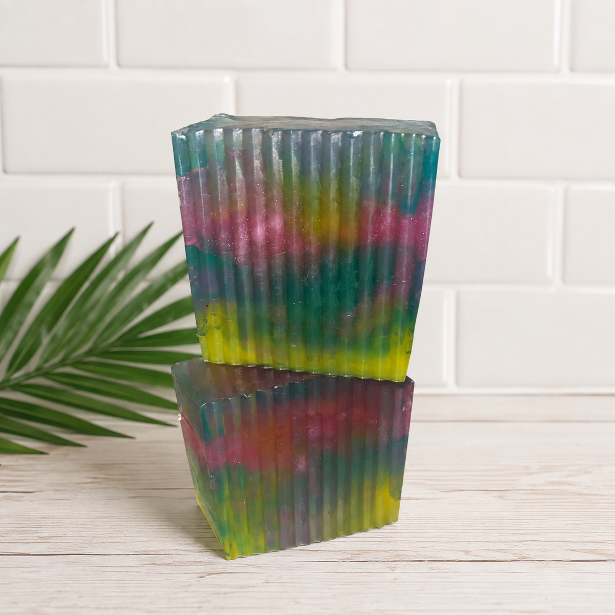 Two translucent pleated soaps stacked on a light wood surface, featuring a multicolor tie-dye pattern of green, pink, yellow, and purple, set against a white subway tile wall with a green palm leaf on the left.