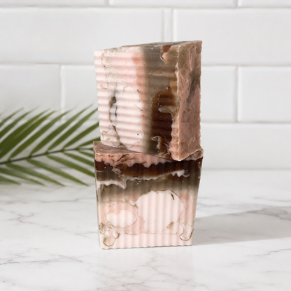 Two stacked rectangular bars of handmade soap with a marbled pattern of pink, brown, and cream, standing on a white marble surface with a white subway tile wall and a green palm leaf in the background.
