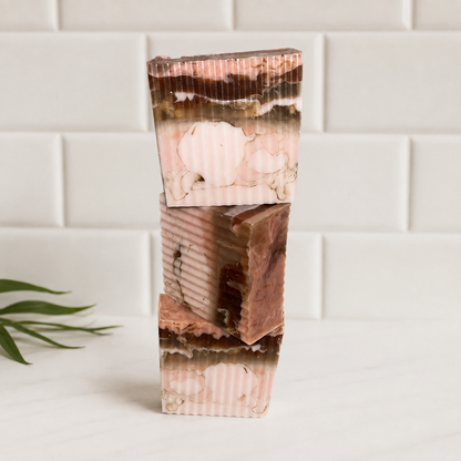 Three stacked rectangular bars of layered soap with pink, brown, and cream marbled patterns, standing on a white marble surface against a white subway tile background.