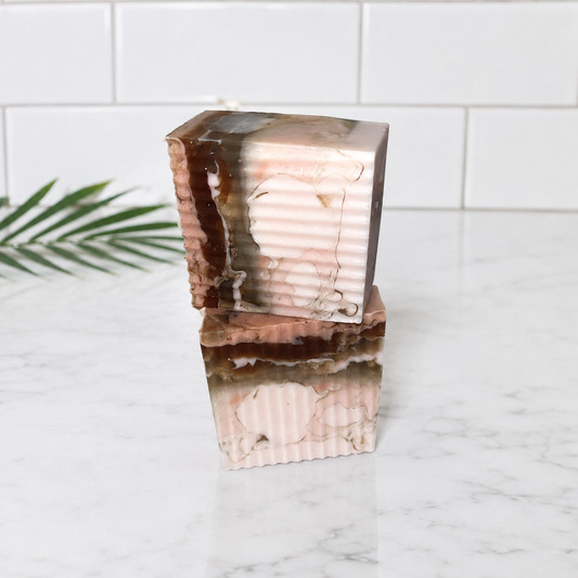 Two stacked rectangular bars of handmade soap with a pink, brown, and white marble pattern, set on a white marble countertop with a white subway tile backsplash.