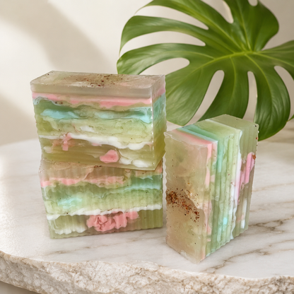 Three layered translucent bars of soap with pink, mint green, and white marbling, displayed on a marble slab with a green monstera leaf in the background.