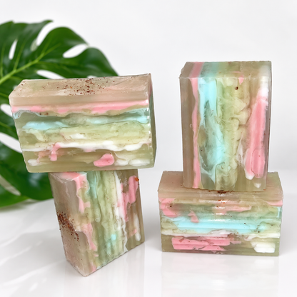 Three rectangular handmade soap bars with marbled pastel layers of pink, mint green, beige, and white, displayed on a white surface with a green leaf in the background.