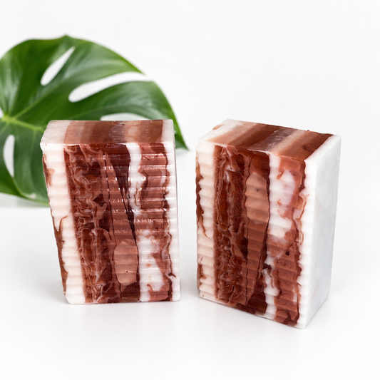 Two rectangular bars of handmade soap with a marbled pattern of dark reddish-brown and white, displayed on a white surface with a green monstera leaf in the background.