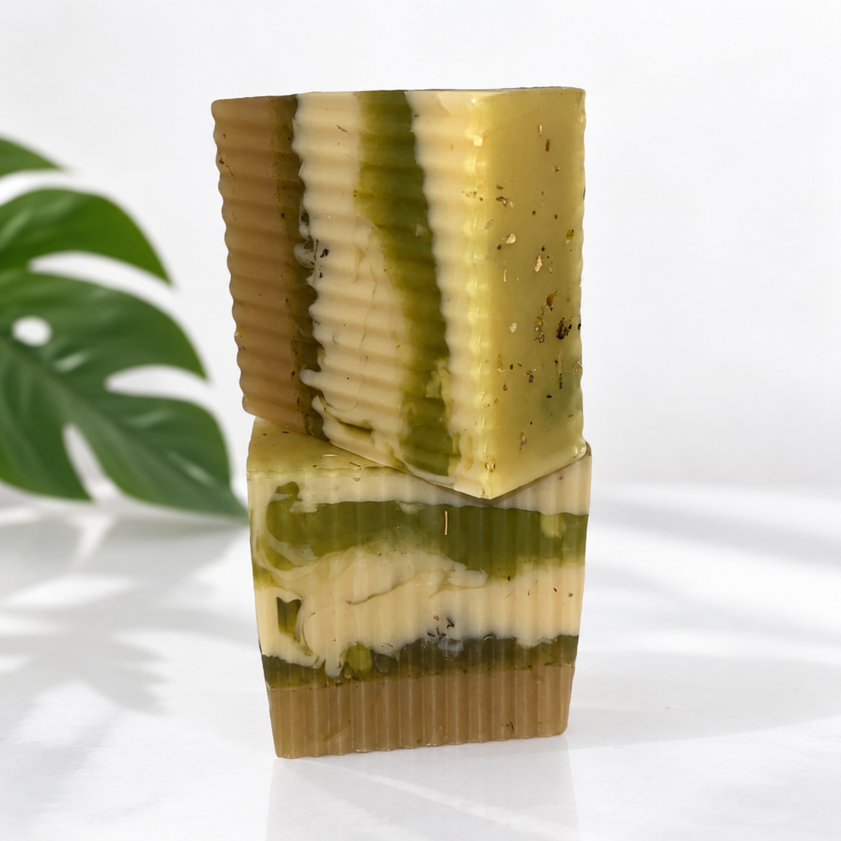 Two stacked rectangular bars of handmade soap with a marbled pattern of olive green, cream, and tan, set against a white background with a green monstera leaf on the left.