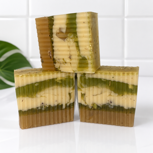 Three rectangular handmade soap bars stacked in a pyramid arrangement on a white surface, with a white tiled wall and a green leaf in the background. The soaps have horizontal layers of olive green, cream, and tan.